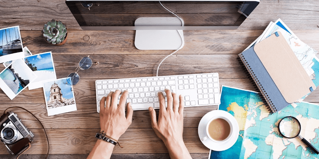 Hands at a keyboard typing on a desk scattered with pictures, camera, tape, plants, maps, magnify glass and sunglasses